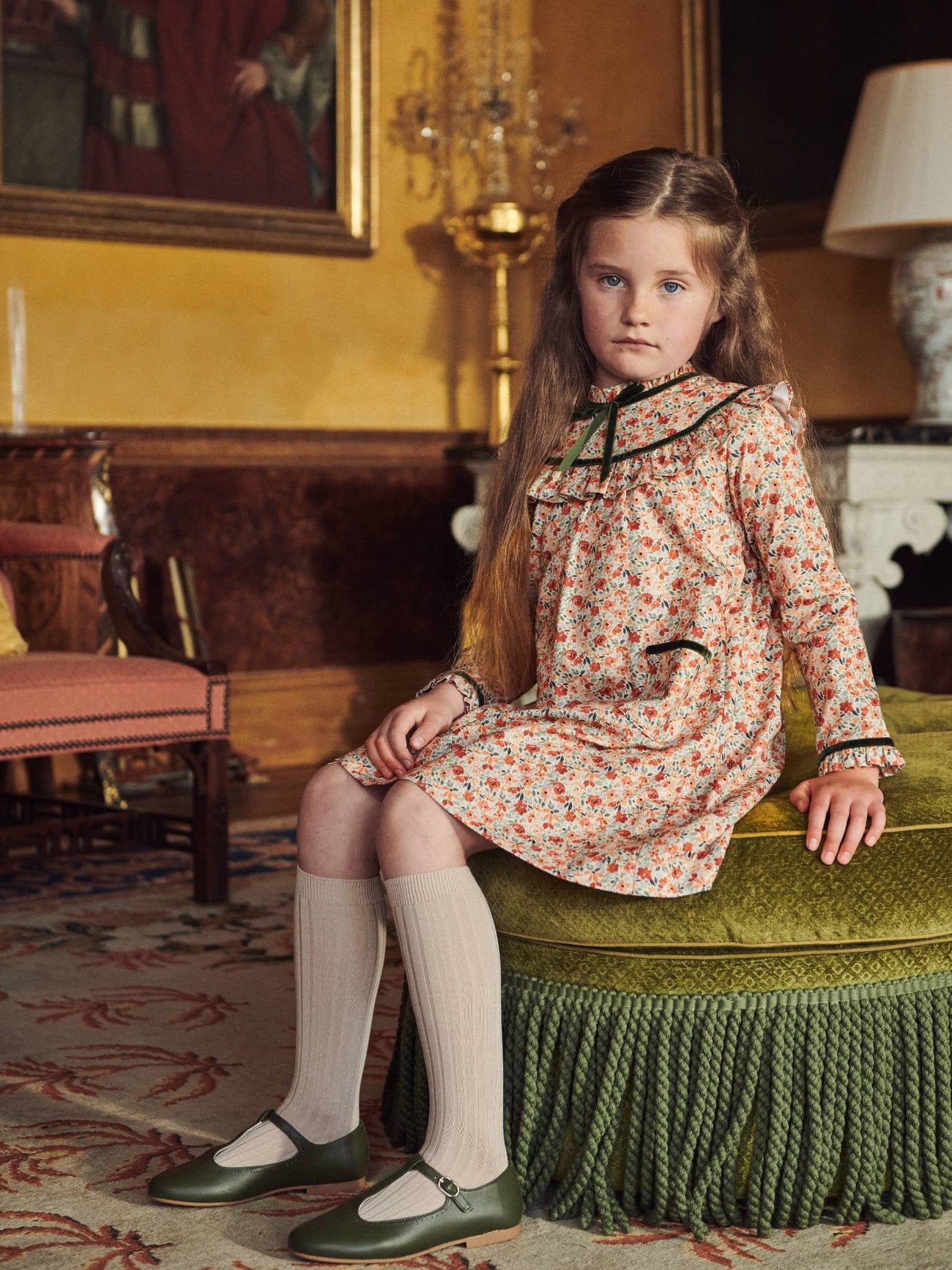 A young girl in an elegant floral print dress with green accents, seated on a fringed ottoman, in a classic, warm room.