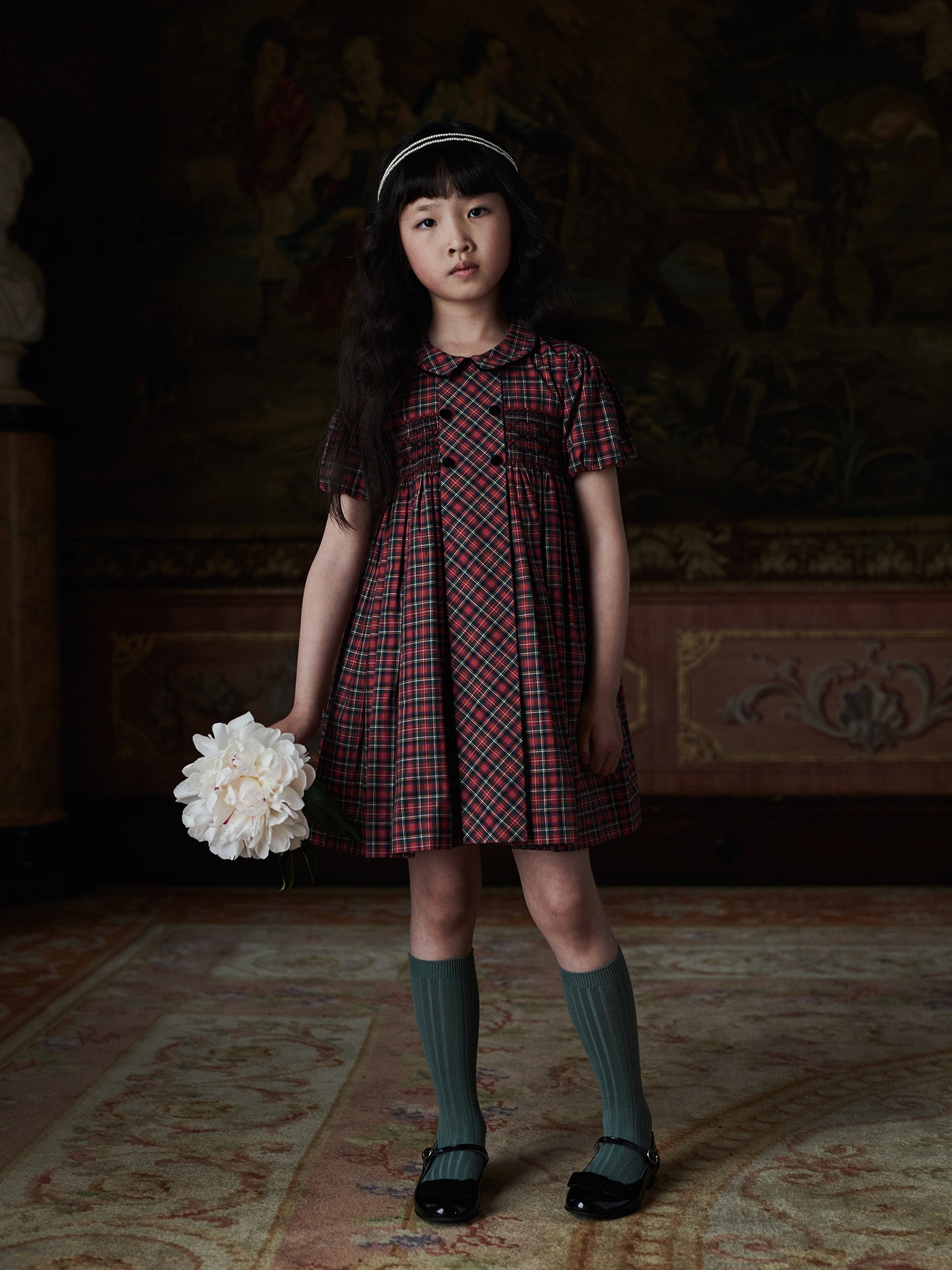 A young girl in a stylish red and black plaid dress with a Peter Pan collar, dark green knee-high socks, and black Mary Jane shoes.