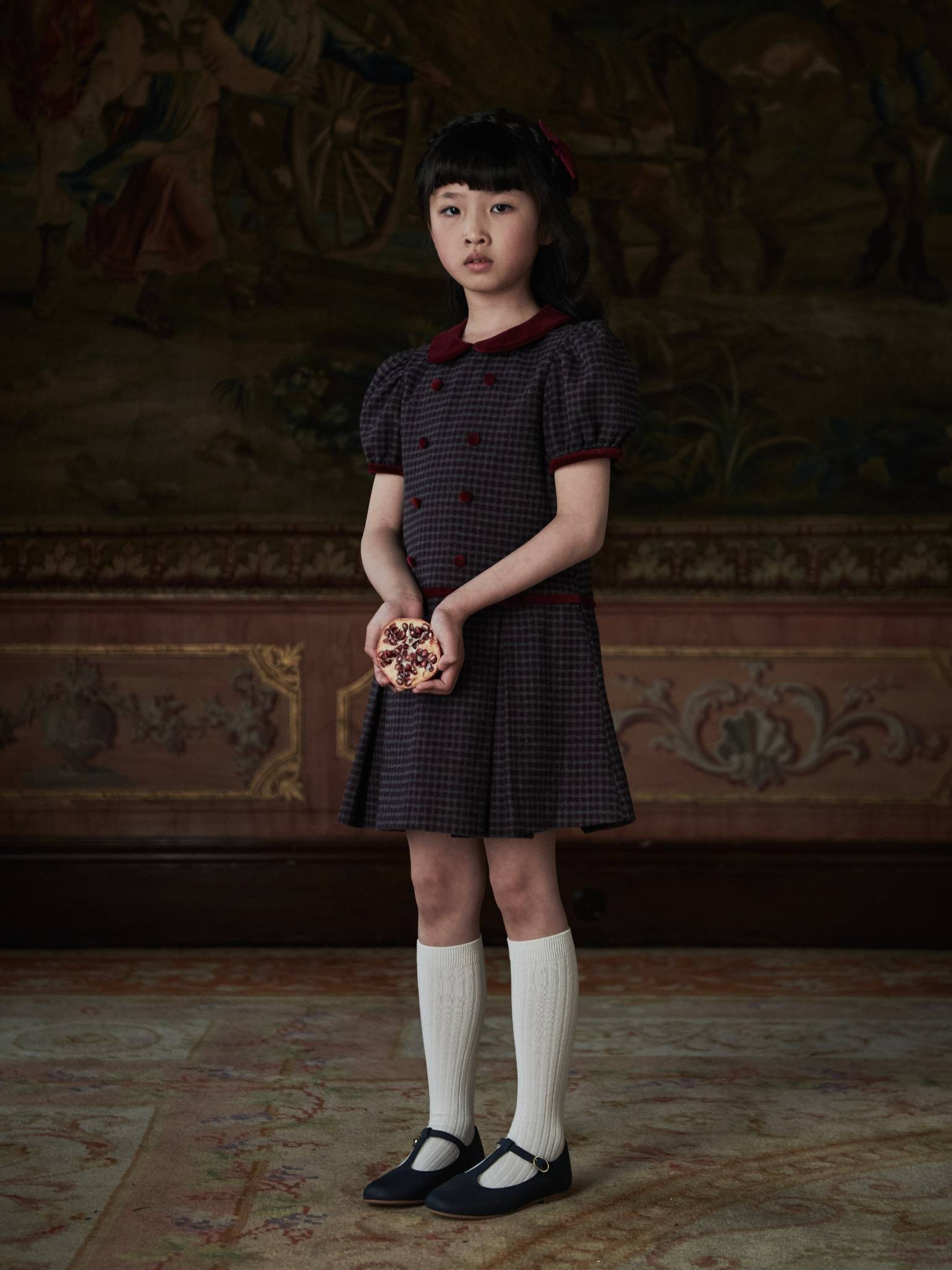 Young girl in elegant children’s fashion, wearing a dark plaid dress with red accents, white knee-high socks, and black Mary Jane shoes.