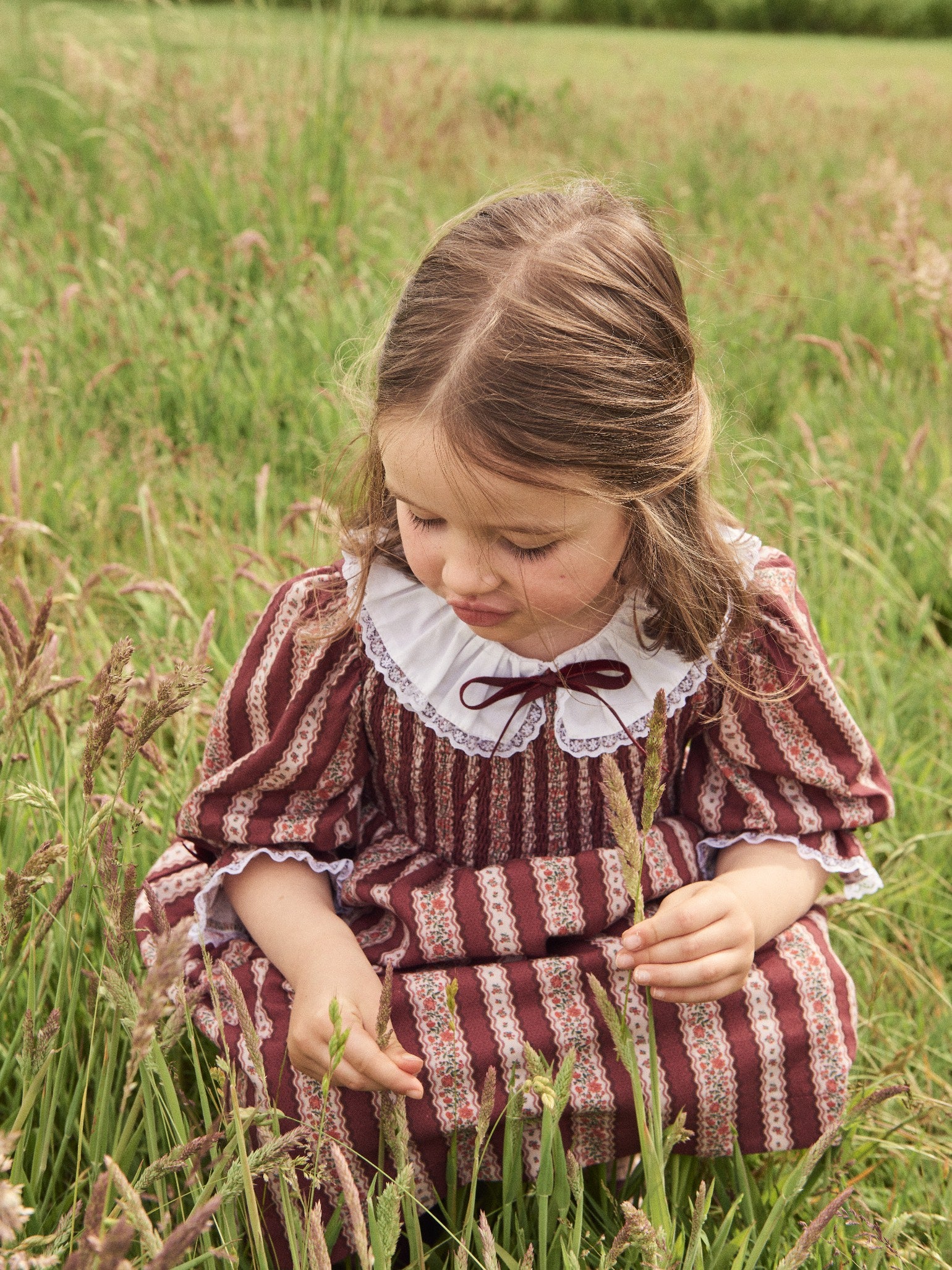 Young girl in a vintage brown striped dress with lace collar sits gracefully in tall grass, exploring and observing nature.