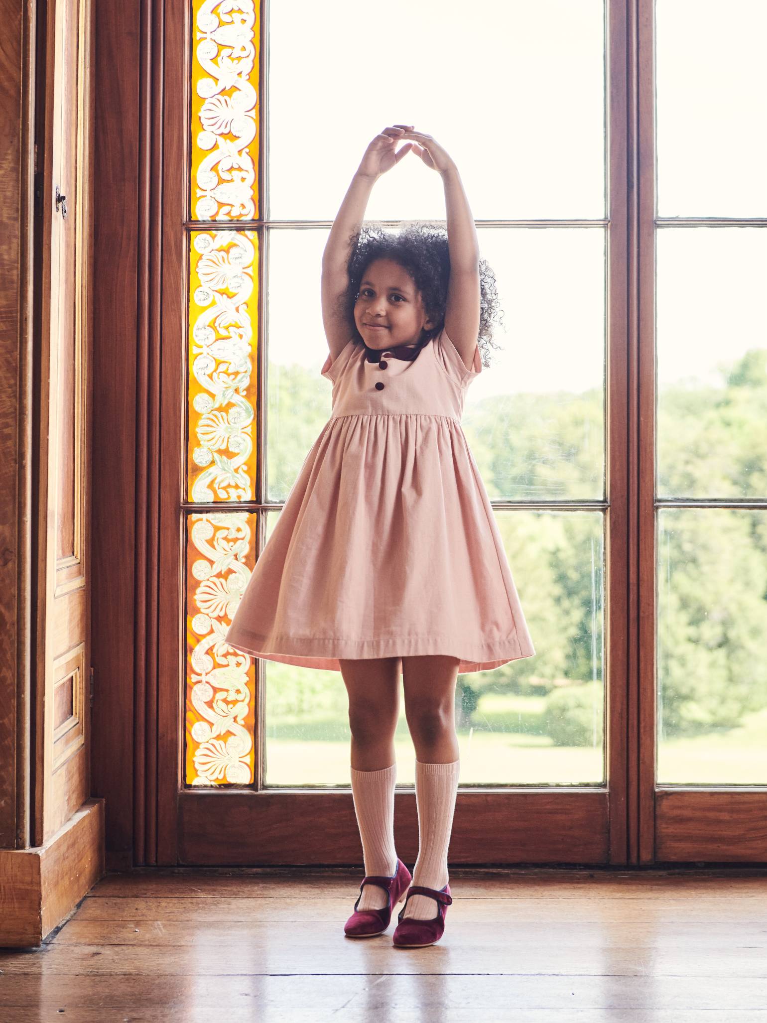 A young girl poses gracefully in a charming light pink dress, paired with white knee-high socks and vibrant red Mary Jane shoes.