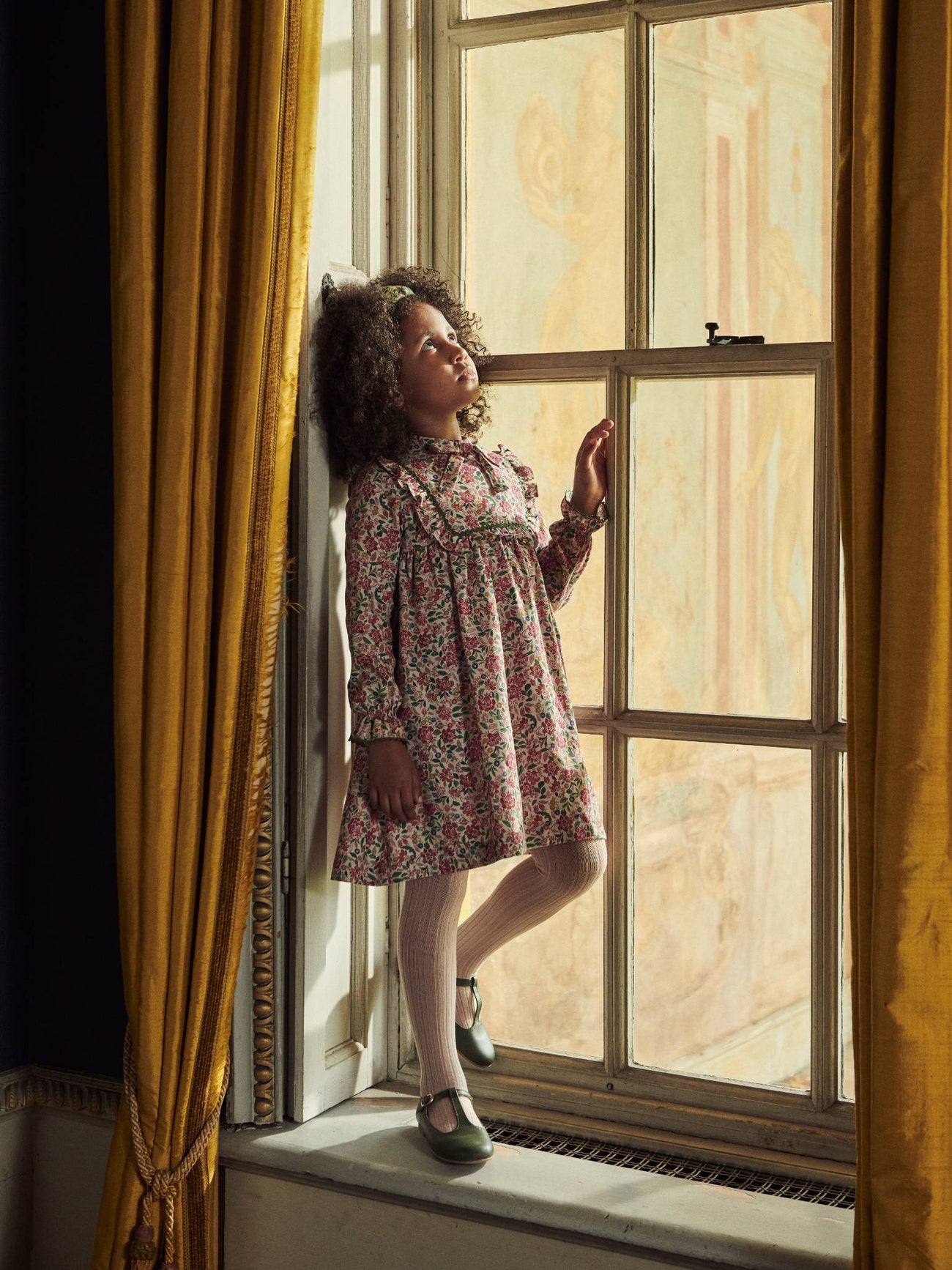A young girl gazes out a window, wearing a floral dress with a ruffled collar, beige tights, and stylish dark green Mary Jane shoes.