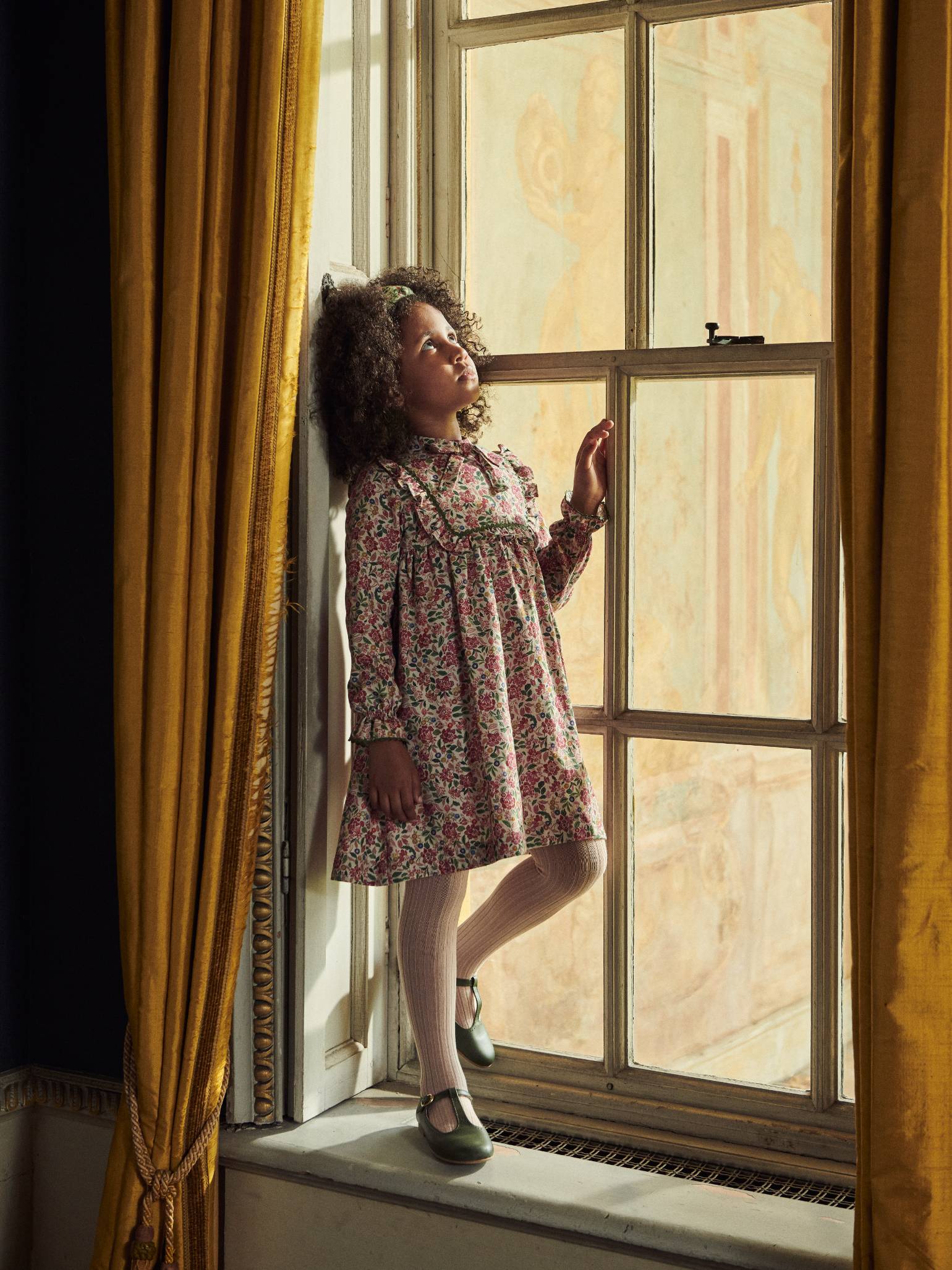 A young girl gazes out a window, wearing a floral dress with a ruffled collar, beige tights, and stylish dark green Mary Jane shoes.