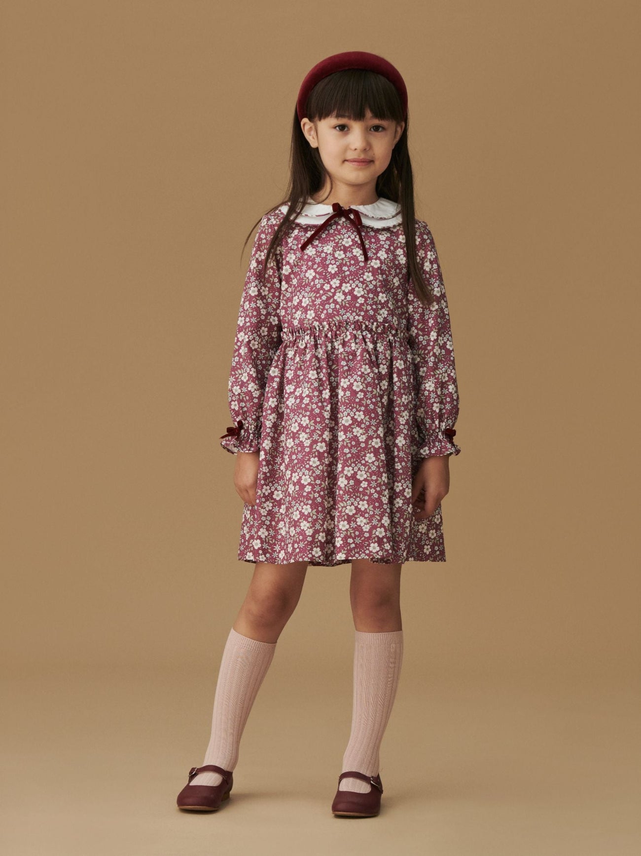 A young girl wears a charming burgundy floral dress with a white Peter Pan collar, paired with pink knee-high socks and burgundy shoes.