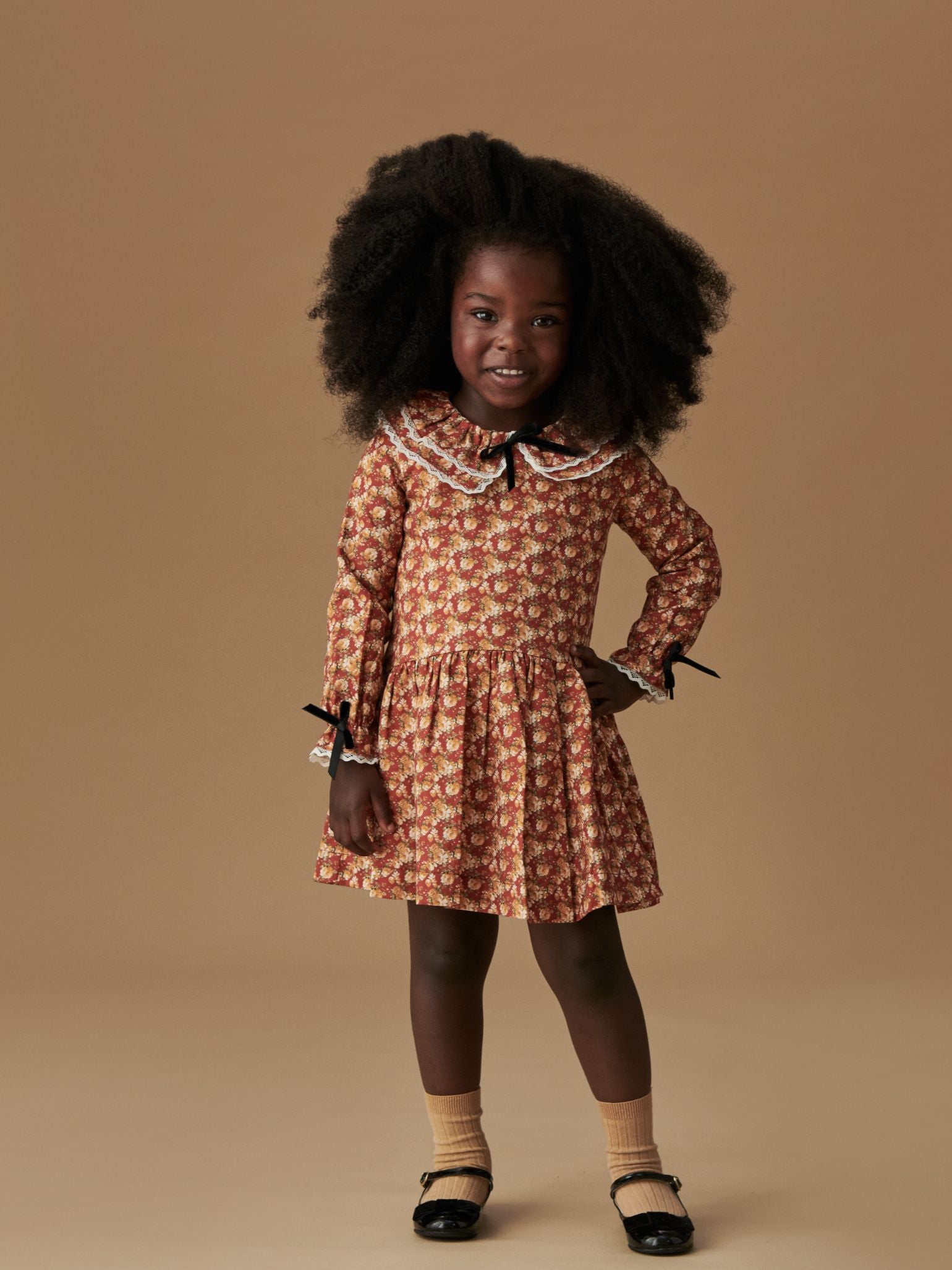 Child models a charming autumn floral dress with lace-trimmed collar, paired with beige socks and classic black Mary Jane shoes.