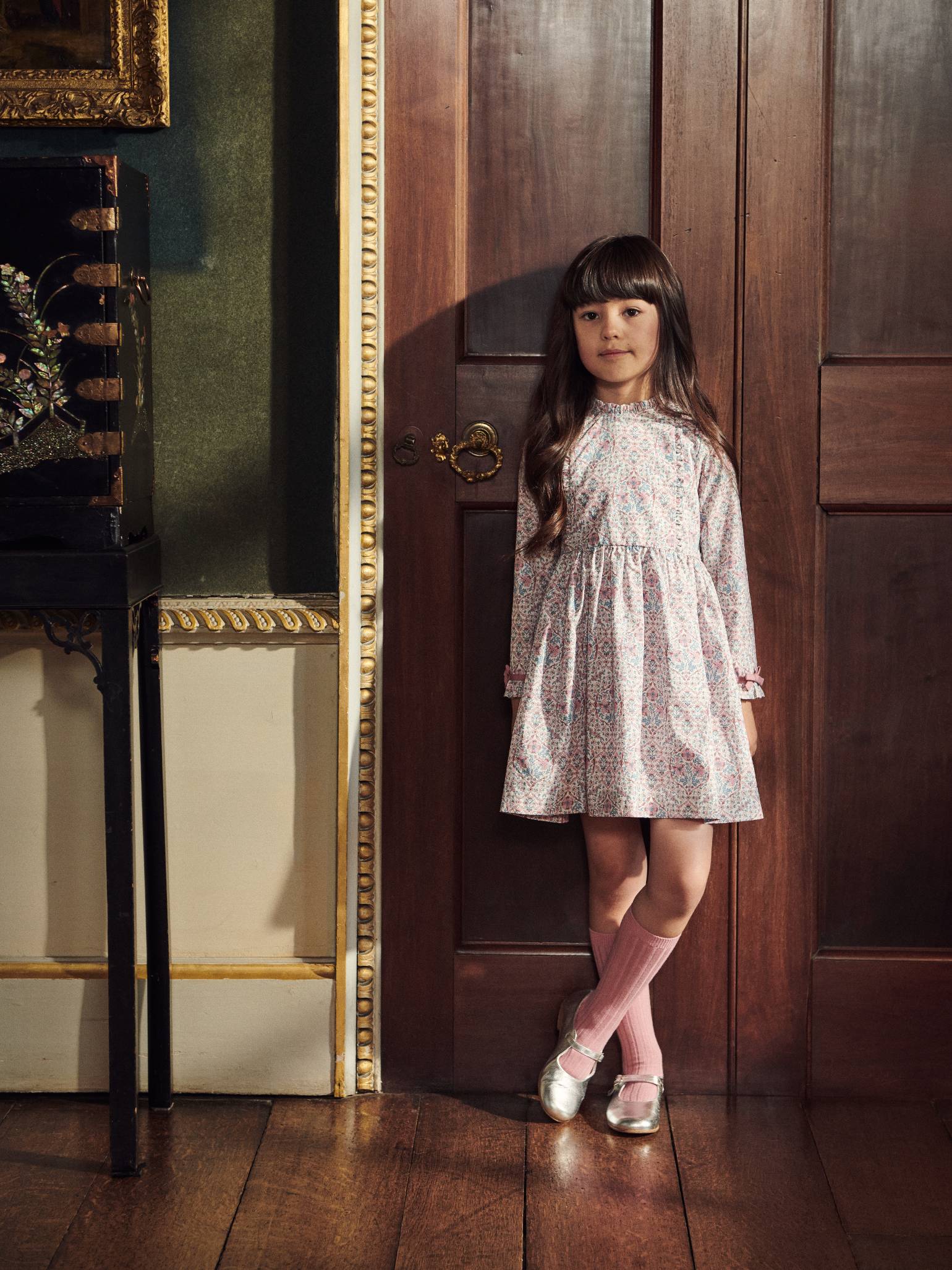 A young girl models a charming floral print dress with long sleeves, complemented by pink knee-high socks and metallic shoes.