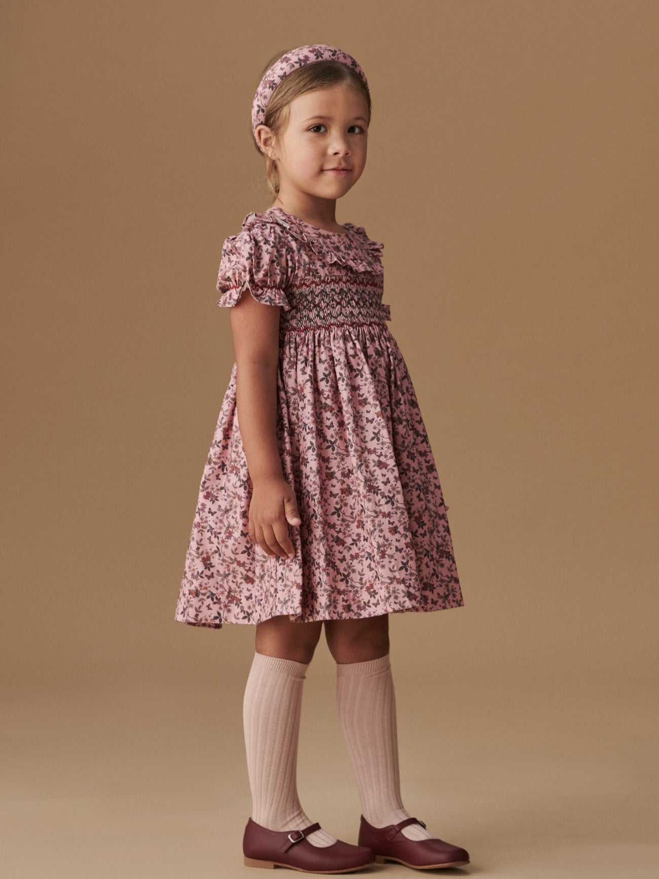 A young girl models a charming pink floral dress with puff sleeves, paired with pink knee-high socks and maroon Mary Jane shoes.