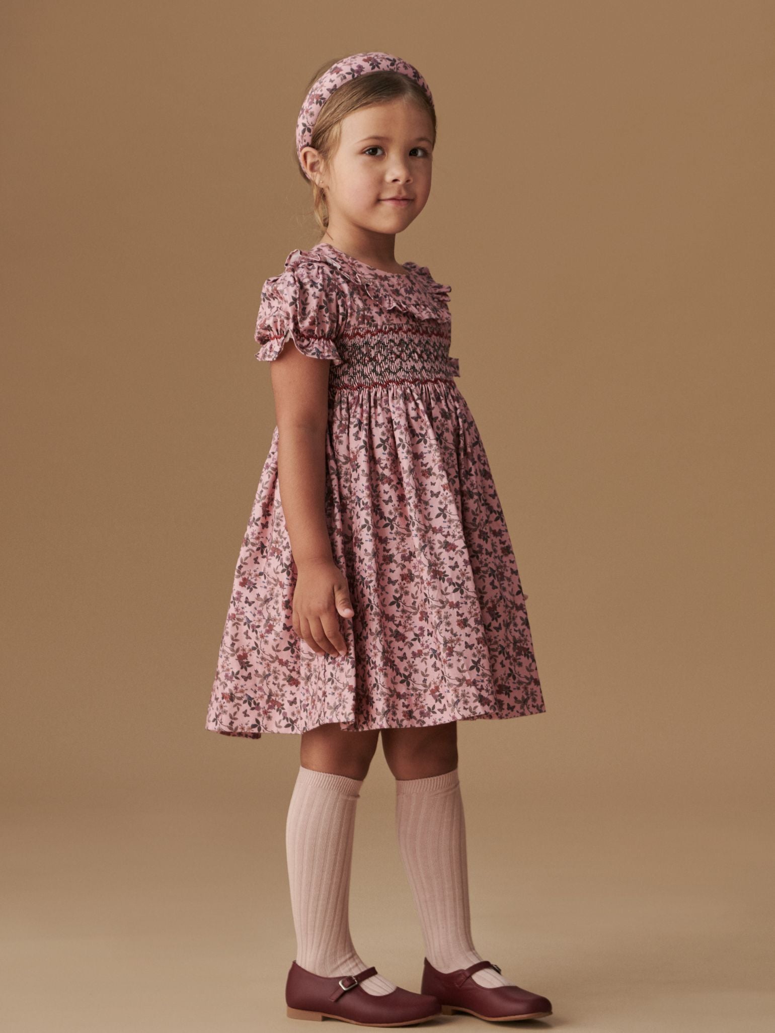 A young girl models a charming pink floral dress with puff sleeves, paired with pink knee-high socks and maroon Mary Jane shoes.