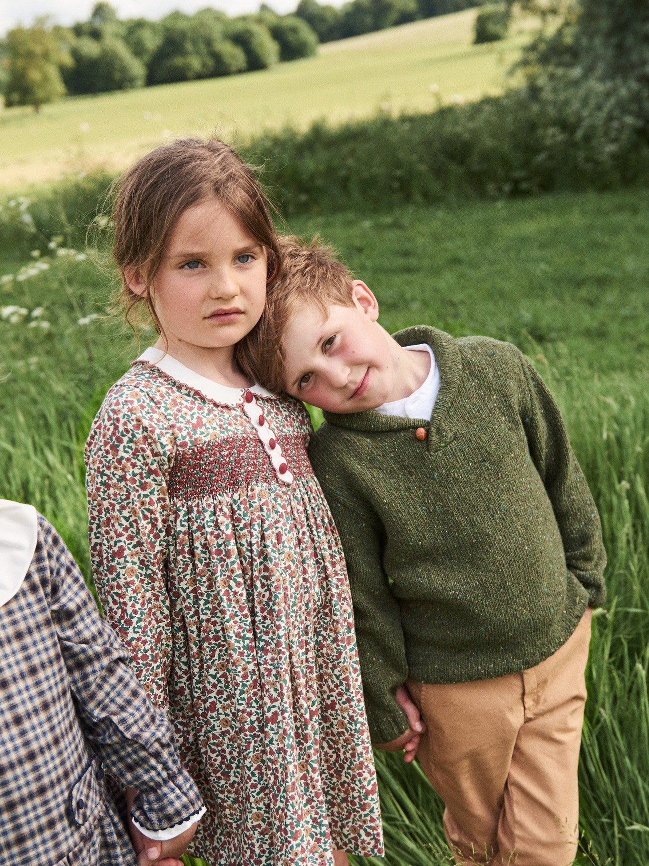 A girl in a floral dress with a Peter Pan collar and a boy in a green sweater stand together in a grassy field.