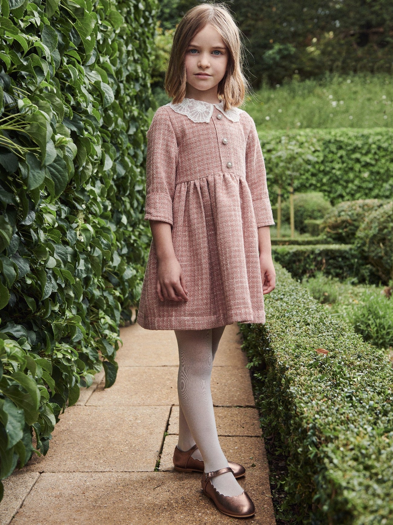 A young girl elegantly models a pink tweed dress with lace collar, cream tights, and bronze Mary Jane shoes in a lush garden setting.