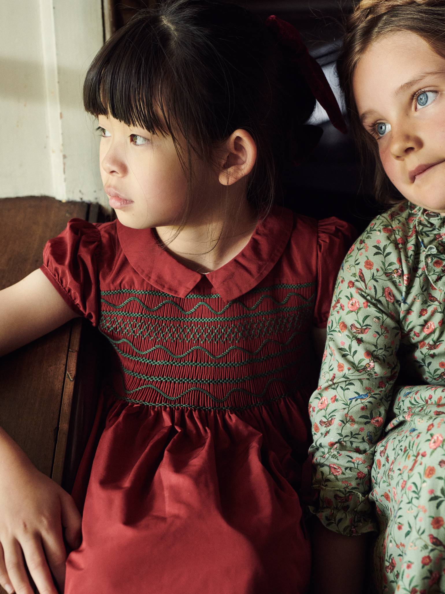 Two young girls in vintage-style dresses, one in red with embroidery, the other in floral green, sitting by a window.