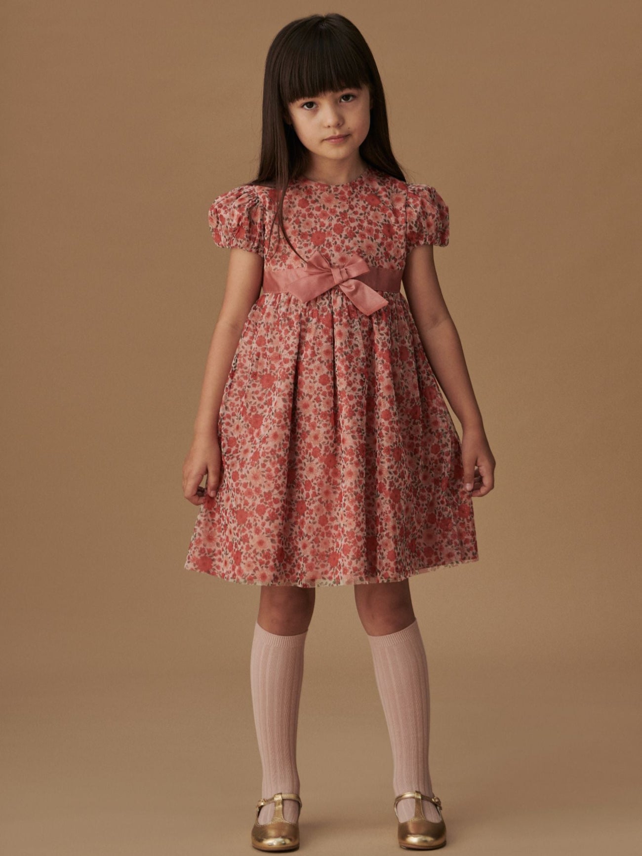 A young girl models an elegant floral children's dress in shades of red and pink with a bow, paired with gold shoes and pink knee-high socks.