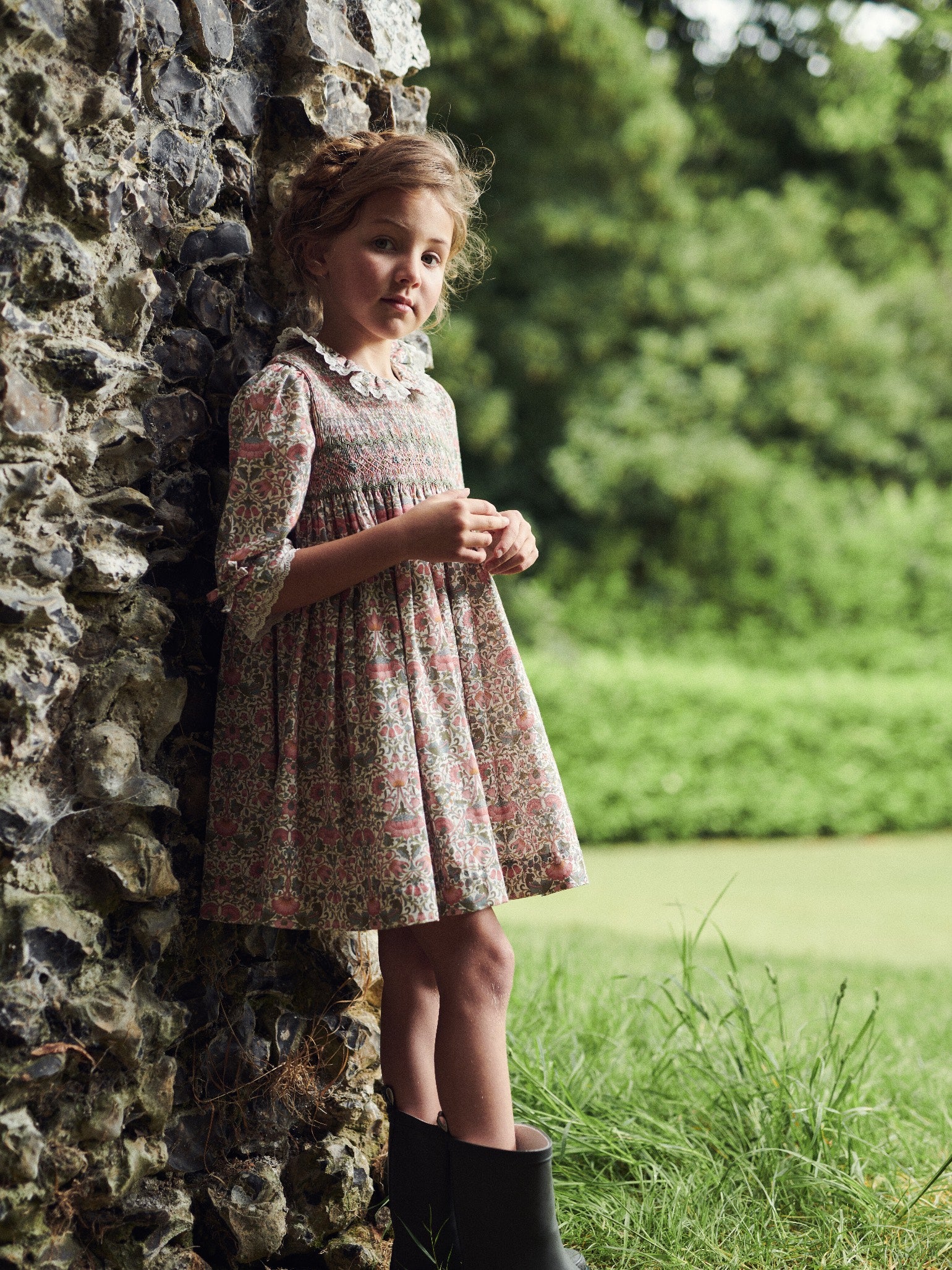 A young girl in a vintage floral dress with lace collar, standing thoughtfully beside a rustic stone wall in a lush garden setting.
